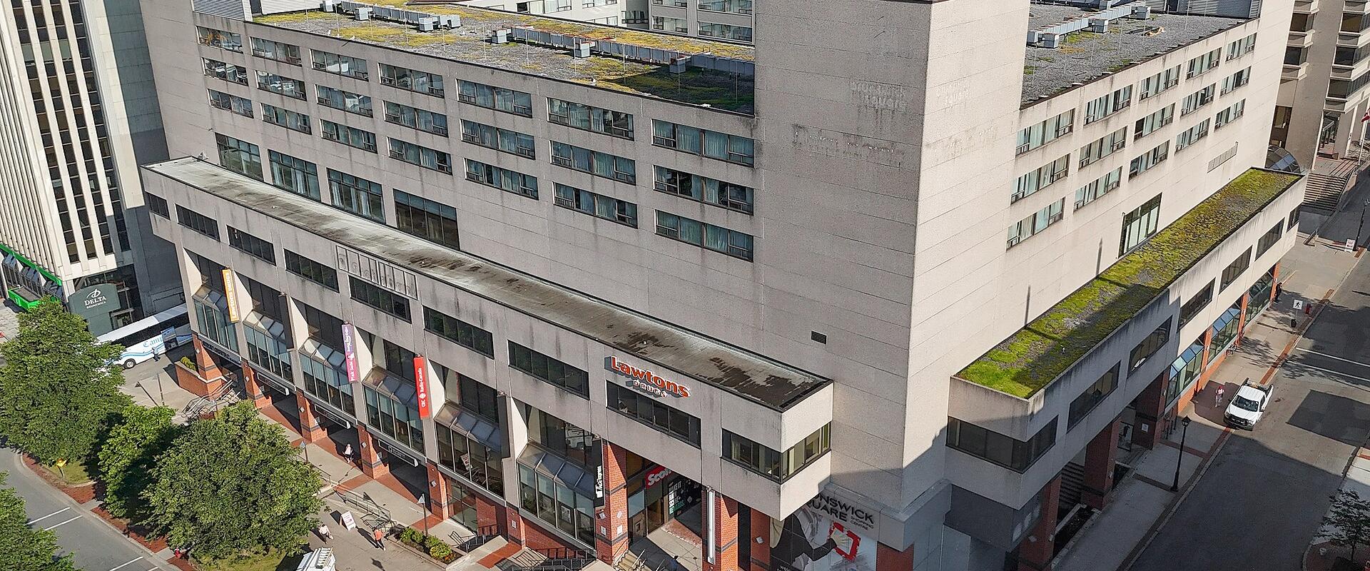 An aerial view of a large, multi-story commercial building complex featuring a "Lawtons Drugs" storefront and a "New Brunswick" banner on the facade, situated in an urban downtown area.