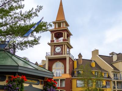 A pedestrian village square featuring a prominent clock tower with a copper-colored spire, surrounded by colorful European-style buildings and an information kiosk in the foreground.