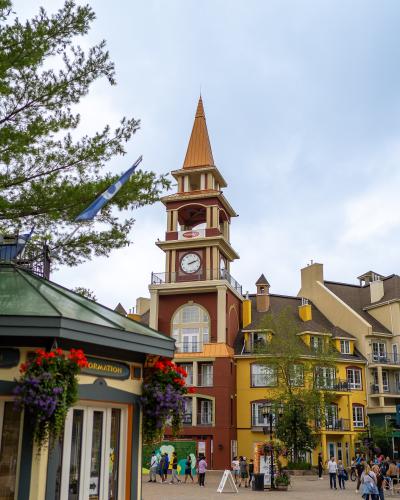 A pedestrian village square featuring a prominent clock tower with a copper-colored spire, surrounded by colorful European-style buildings and an information kiosk in the foreground.
