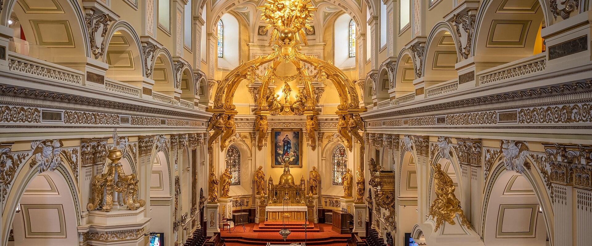 The image shows the ornate interior of a large church, featuring a prominent golden baldachin over the main altar, white walls with gold trim, and a high vaulted ceiling.
