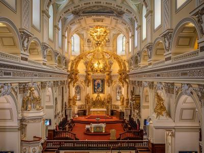 The image shows the ornate interior of a large church, featuring a prominent golden baldachin over the main altar, white walls with gold trim, and a high vaulted ceiling.