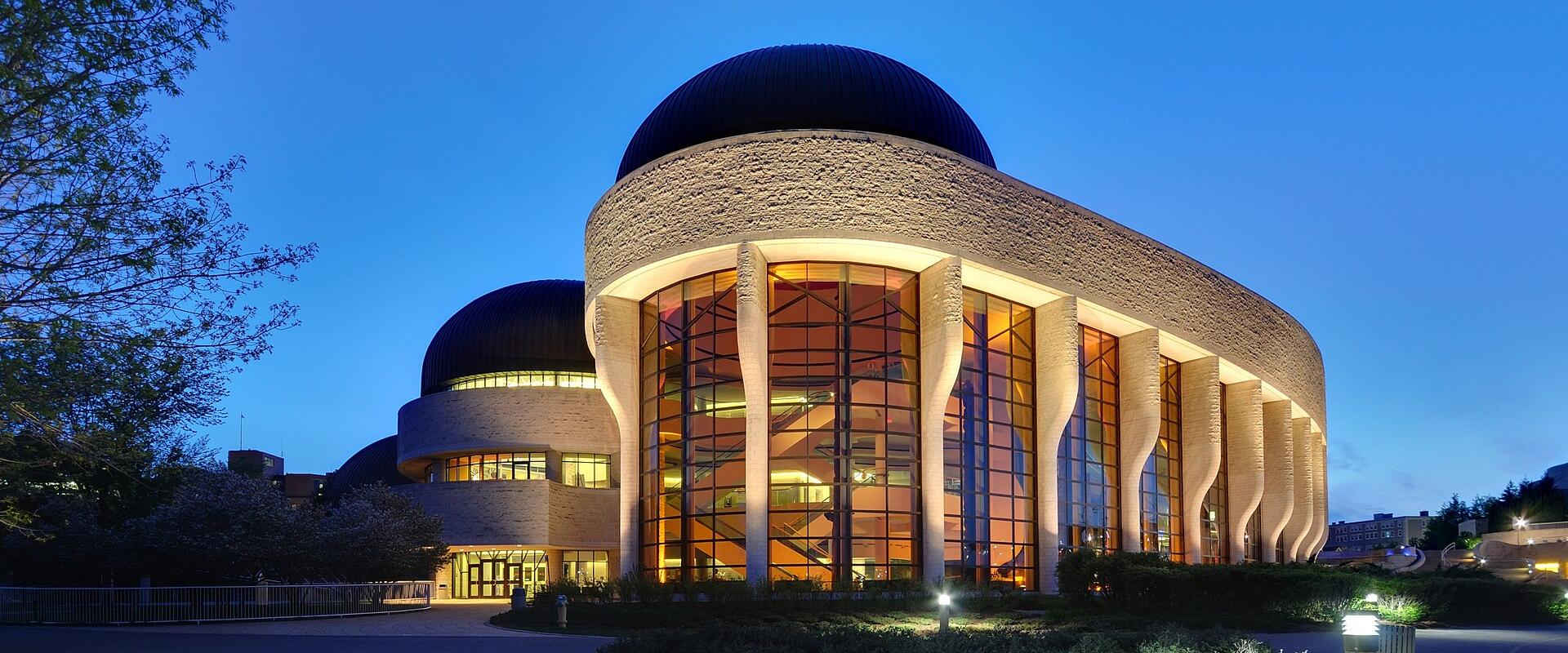 A large, modern building with distinctive curved stone walls, massive glass windows, and dark domes, illuminated at twilight.