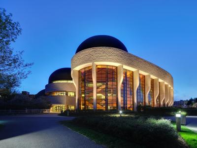 A large, modern building with distinctive curved stone walls, massive glass windows, and dark domes, illuminated at twilight.