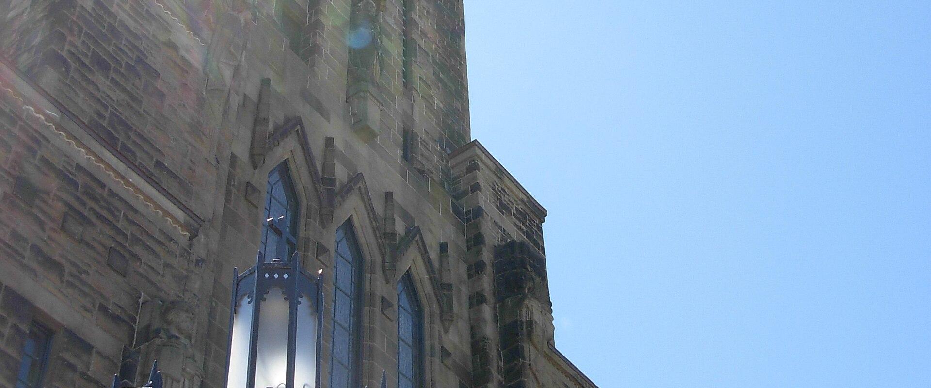 A low-angle view of a tall, stone cathedral tower with Gothic Revival architectural features, including pointed arch windows and vertical buttresses, with a decorative blue lamppost in the foreground.