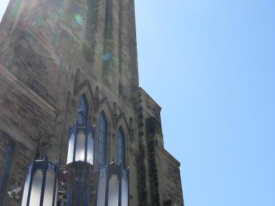 A low-angle view of a tall, stone cathedral tower with Gothic Revival architectural features, including pointed arch windows and vertical buttresses, with a decorative blue lamppost in the foreground.