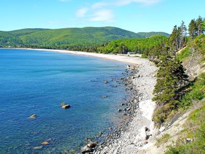 A scenic coastal view showing a long sandy beach curving along a bay, bordered by a rocky shoreline and steep, forested cliffs in the foreground, with green hills in the background under a blue sky.