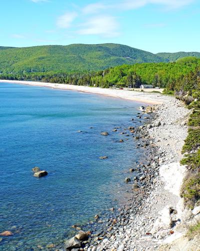 A scenic coastal view showing a long sandy beach curving along a bay, bordered by a rocky shoreline and steep, forested cliffs in the foreground, with green hills in the background under a blue sky.