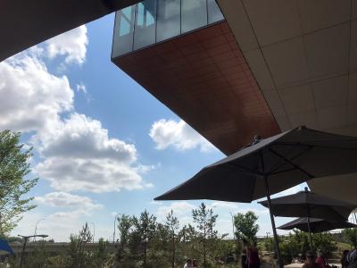 A low-angle view looking up at the cantilevered, copper-paneled underside of a modern building's overhang, with large patio umbrellas and people visible below against a blue sky.