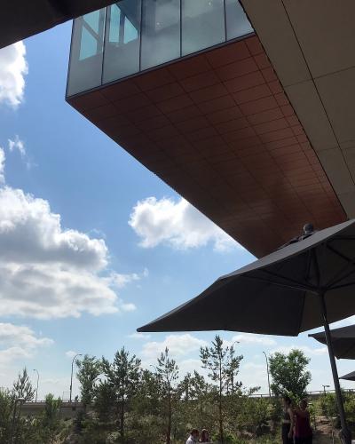 A low-angle view looking up at the cantilevered, copper-paneled underside of a modern building's overhang, with large patio umbrellas and people visible below against a blue sky.