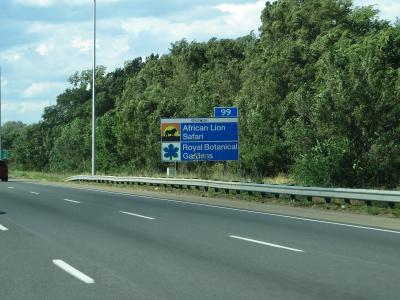 A blue highway exit sign on the Queen Elizabeth Way in Ontario, displaying directions for "African Lion Safari" and "Royal Botanical Gardens" with a leaf logo.