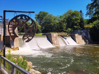 A concrete dam or weir with water cascading over it, featuring a large, rusted metal sculpture of a fish in the foreground and a metal fence along the riverbank.