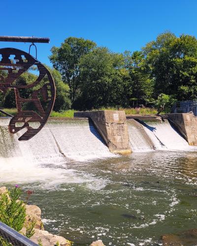 A concrete dam or weir with water cascading over it, featuring a large, rusted metal sculpture of a fish in the foreground and a metal fence along the riverbank.