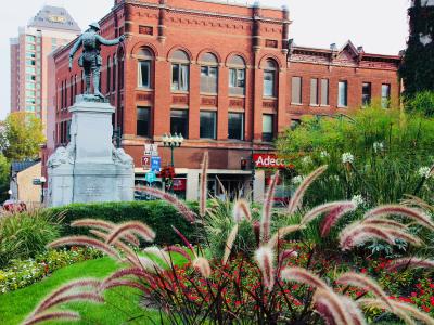 A war memorial statue of a soldier on a stone pedestal stands in a landscaped park area, with red brick commercial buildings (including one with an "Adecco" sign) in the background.