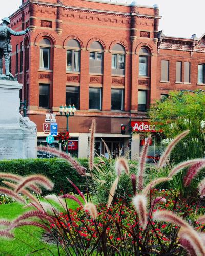 A war memorial statue of a soldier on a stone pedestal stands in a landscaped park area, with red brick commercial buildings (including one with an "Adecco" sign) in the background.