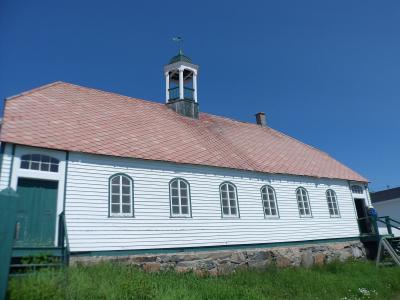 A long, white wooden building with a red shingled roof, green trim, arched windows, and a small cupola topped with a cross, set against a clear blue sky.