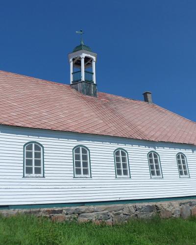 A long, white wooden building with a red shingled roof, green trim, arched windows, and a small cupola topped with a cross, set against a clear blue sky.