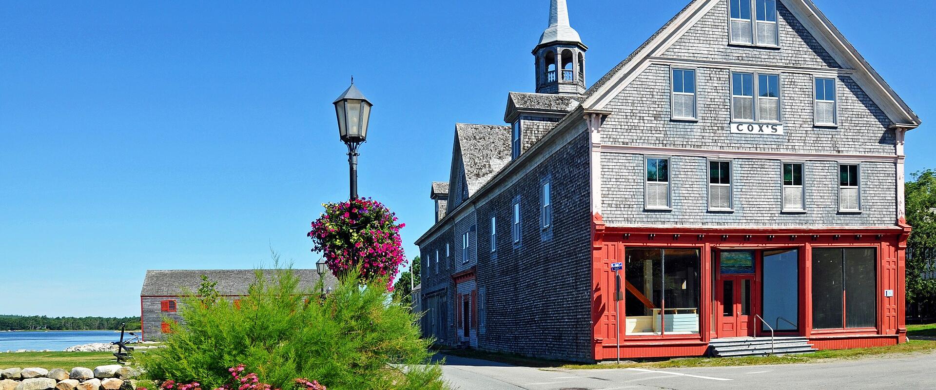 A large, grey-shingled historic building with a red ground-floor facade and a white steeple, situated next to a waterfront with a brick walkway and hanging flower baskets.