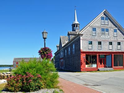 A large, grey-shingled historic building with a red ground-floor facade and a white steeple, situated next to a waterfront with a brick walkway and hanging flower baskets.