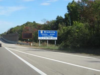 A blue highway welcome sign reading "Brockville, Population 22 000, 3 Interchanges, City of the 1000 Islands" situated on the shoulder of a paved road with trees and a sound barrier in the background.