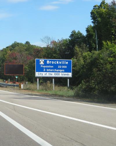 A blue highway welcome sign reading "Brockville, Population 22 000, 3 Interchanges, City of the 1000 Islands" situated on the shoulder of a paved road with trees and a sound barrier in the background.