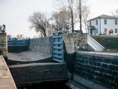 A stone lock chamber with blue metal gates, featuring a white two-story building on the grassy bank to the right and a statue on a pedestal to the left.