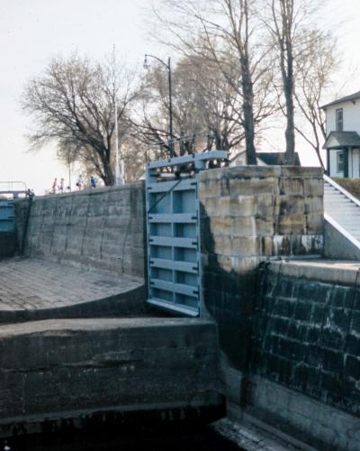 A stone lock chamber with blue metal gates, featuring a white two-story building on the grassy bank to the right and a statue on a pedestal to the left.