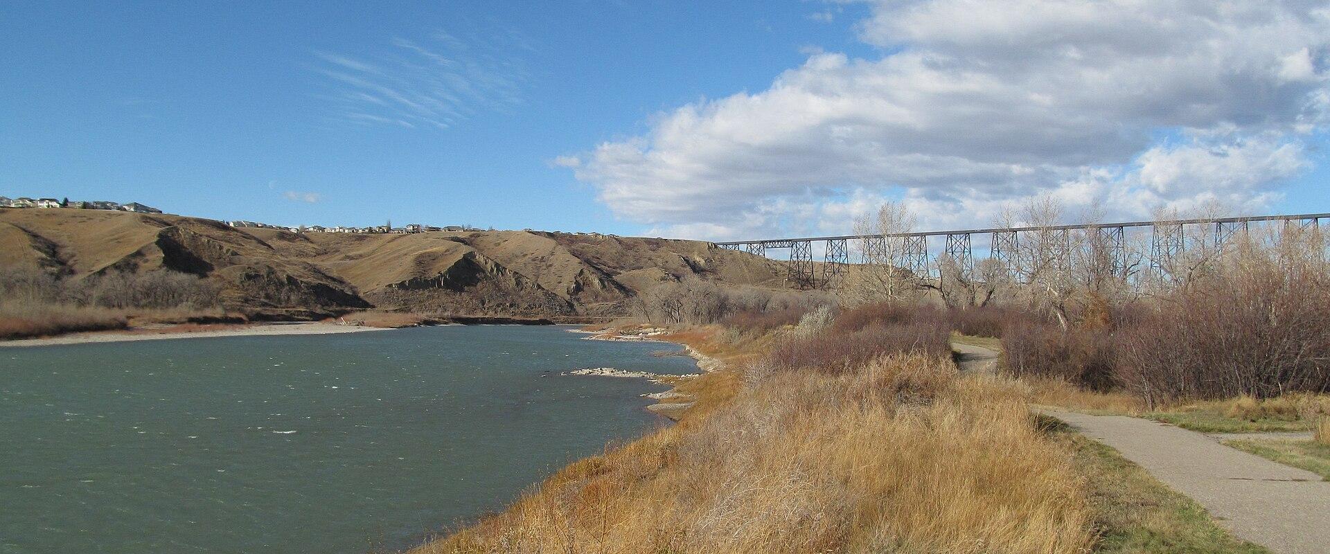 A wide river flows through a valley with steep, grassy banks on the left and a paved pathway along the right bank, with a large steel truss bridge spanning the river in the distance under a blue sky.