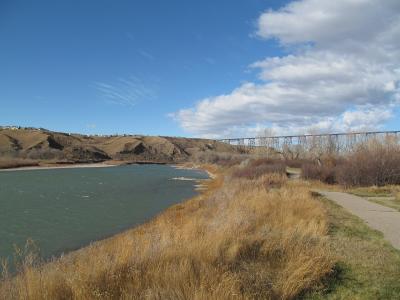 A wide river flows through a valley with steep, grassy banks on the left and a paved pathway along the right bank, with a large steel truss bridge spanning the river in the distance under a blue sky.