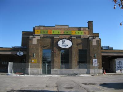 The image shows the exterior facade of a brick building with a colorful sign reading "MARCHÉ JEAN-TALON" and a circular logo for "PREMIÈRE MOISSON" below it, set against a clear blue sky with a fenced-off area in front.