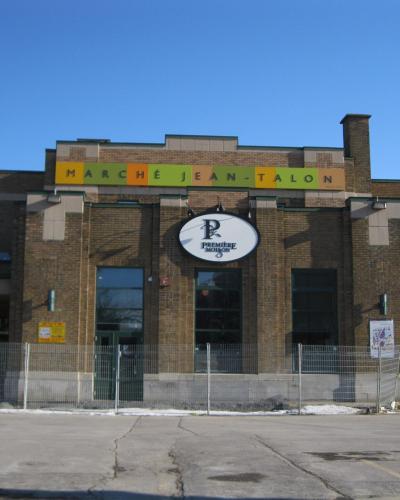 The image shows the exterior facade of a brick building with a colorful sign reading "MARCHÉ JEAN-TALON" and a circular logo for "PREMIÈRE MOISSON" below it, set against a clear blue sky with a fenced-off area in front.