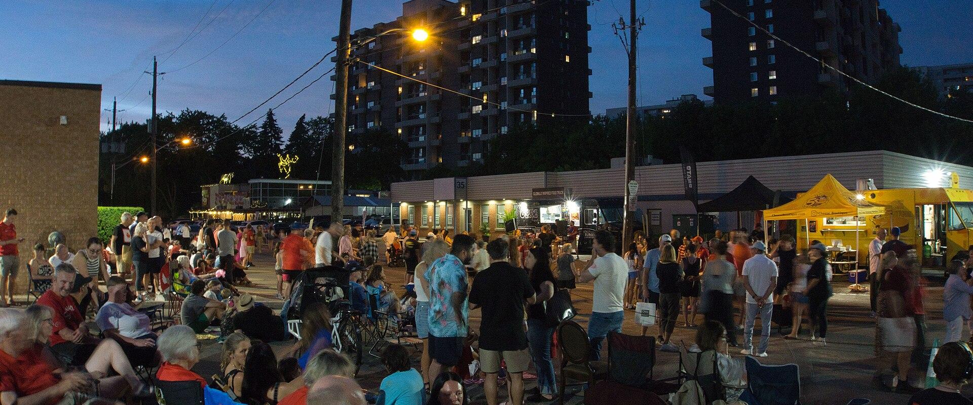 A crowded street festival at dusk with people sitting in lawn chairs, food trucks, and vendor tents set up in front of low-rise commercial buildings and taller apartment blocks.