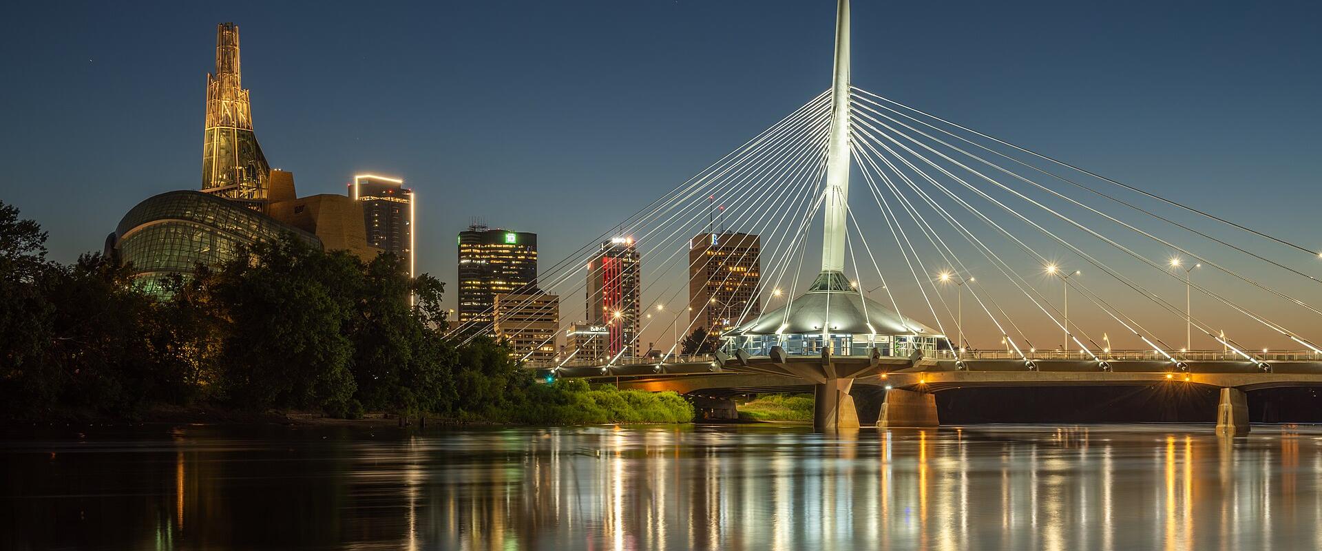 The image shows the Esplanade Riel Bridge, a cable-stayed bridge over the Red River, with the distinctive architecture of The Forks and the Canadian Museum for Human Rights visible in the background skyline at dusk.