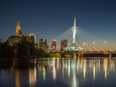 The image shows the Esplanade Riel Bridge, a cable-stayed bridge over the Red River, with the distinctive architecture of The Forks and the Canadian Museum for Human Rights visible in the background skyline at dusk.