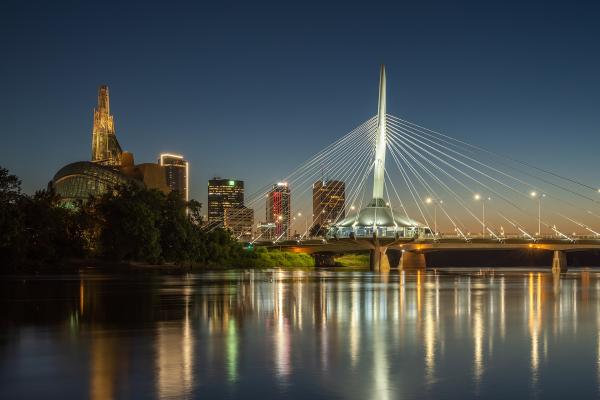 The image shows the Esplanade Riel Bridge, a cable-stayed bridge over the Red River, with the distinctive architecture of The Forks and the Canadian Museum for Human Rights visible in the background skyline at dusk.