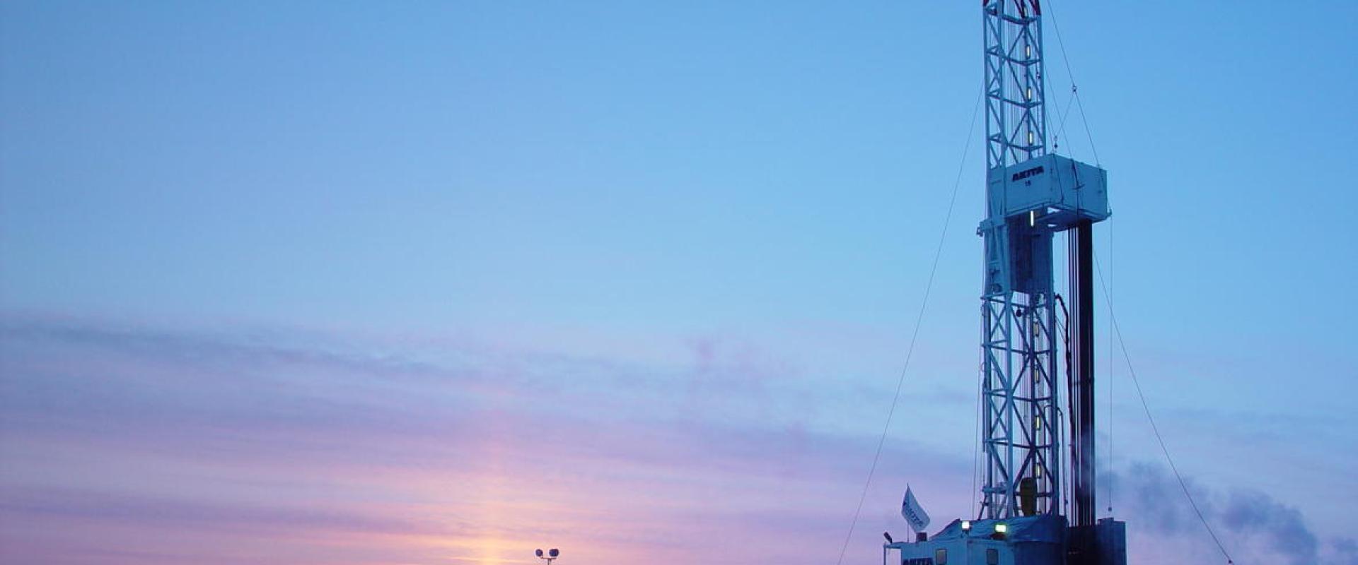 A drilling rig and support equipment situated on a flat, snow-covered landscape during twilight, with a low sun on the horizon.