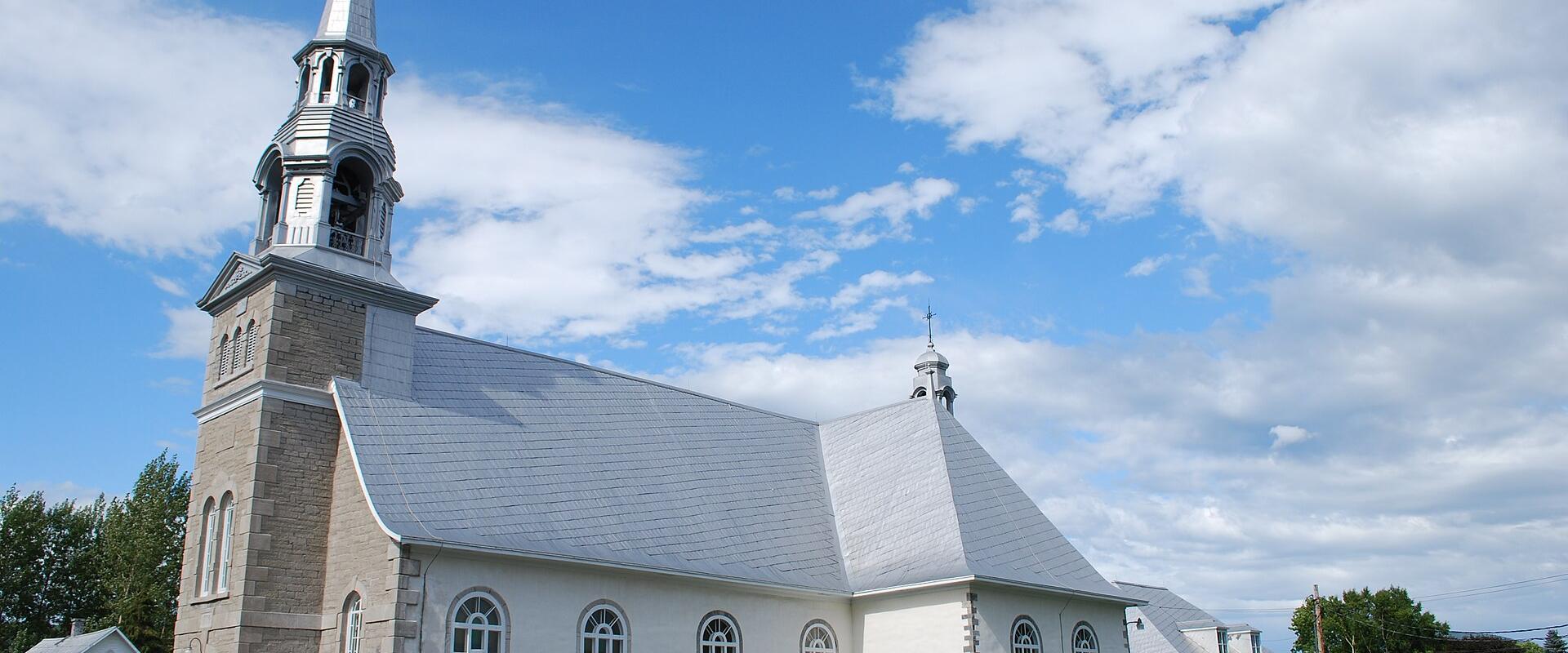A white stone church with a tall bell tower and steeple, set against a blue sky with clouds, with a cemetery visible to the right and a car parked in front.