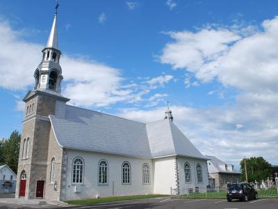 A white stone church with a tall bell tower and steeple, set against a blue sky with clouds, with a cemetery visible to the right and a car parked in front.