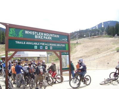 A large green wooden sign reading "WHISTLER MOUNTAIN BIKE PARK" with trail difficulty ratings, situated at the base of a ski hill with chairlifts and riders.