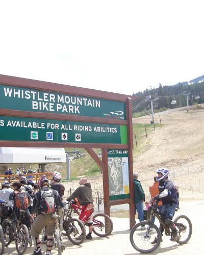 A large green wooden sign reading "WHISTLER MOUNTAIN BIKE PARK" with trail difficulty ratings, situated at the base of a ski hill with chairlifts and riders.