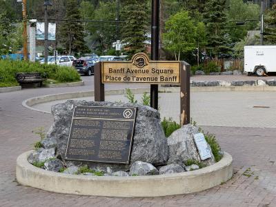 A wooden sign reading "Banff Avenue Square / Place de l'avenue Banff" mounted on a post behind a historical plaque set in rocks within a paved plaza.