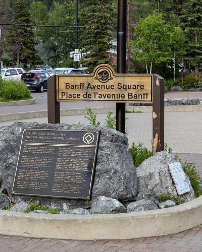 A wooden sign reading "Banff Avenue Square / Place de l'avenue Banff" mounted on a post behind a historical plaque set in rocks within a paved plaza.