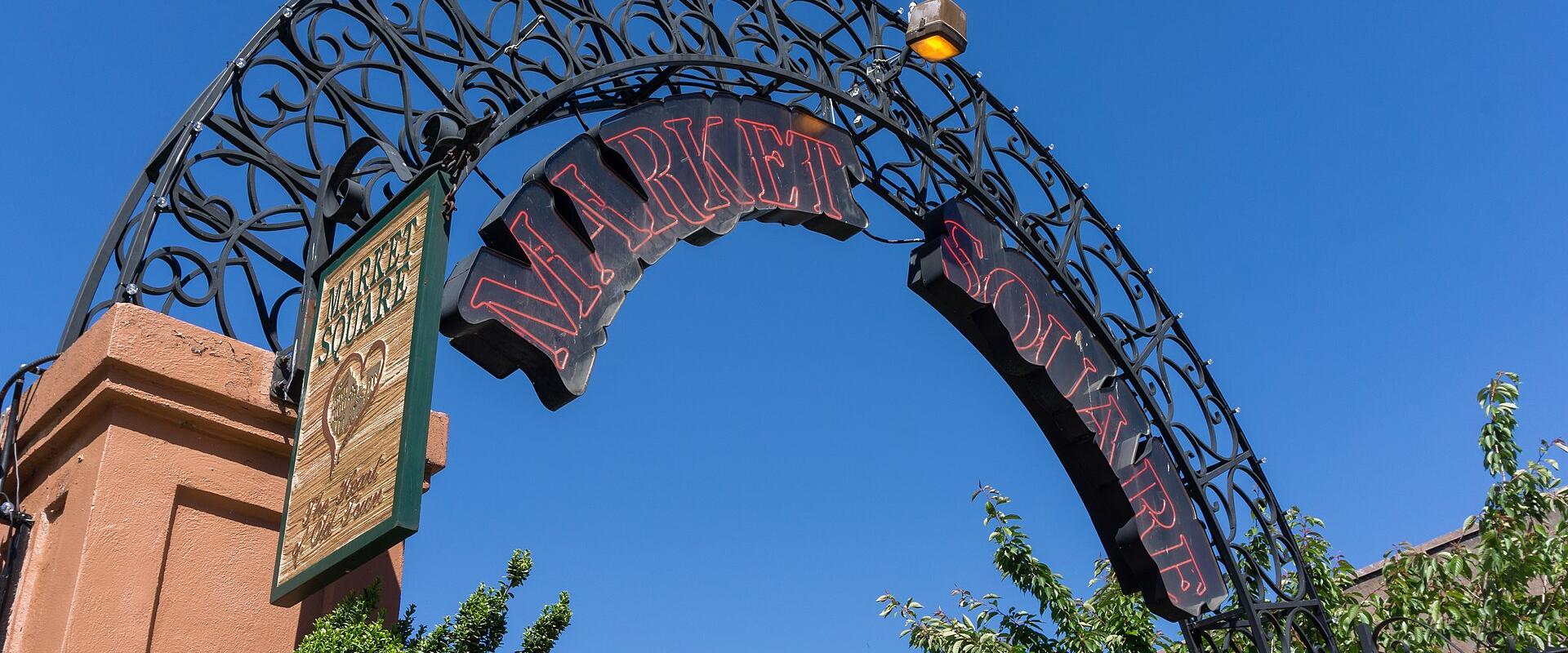 A low-angle shot of an ornate black wrought-iron archway with red neon lettering spelling "MARKET SQUARE" and a wooden sign on the left pillar also reading "MARKET SQUARE".