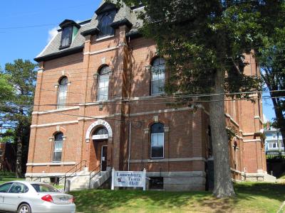 A red brick building with arched windows and a mansard roof, identified by a white sign in the foreground and text above the entrance as the "Lunenburg Town Hall".