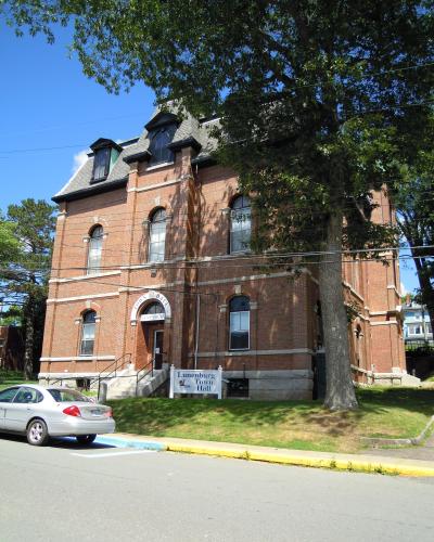 A red brick building with arched windows and a mansard roof, identified by a white sign in the foreground and text above the entrance as the "Lunenburg Town Hall".