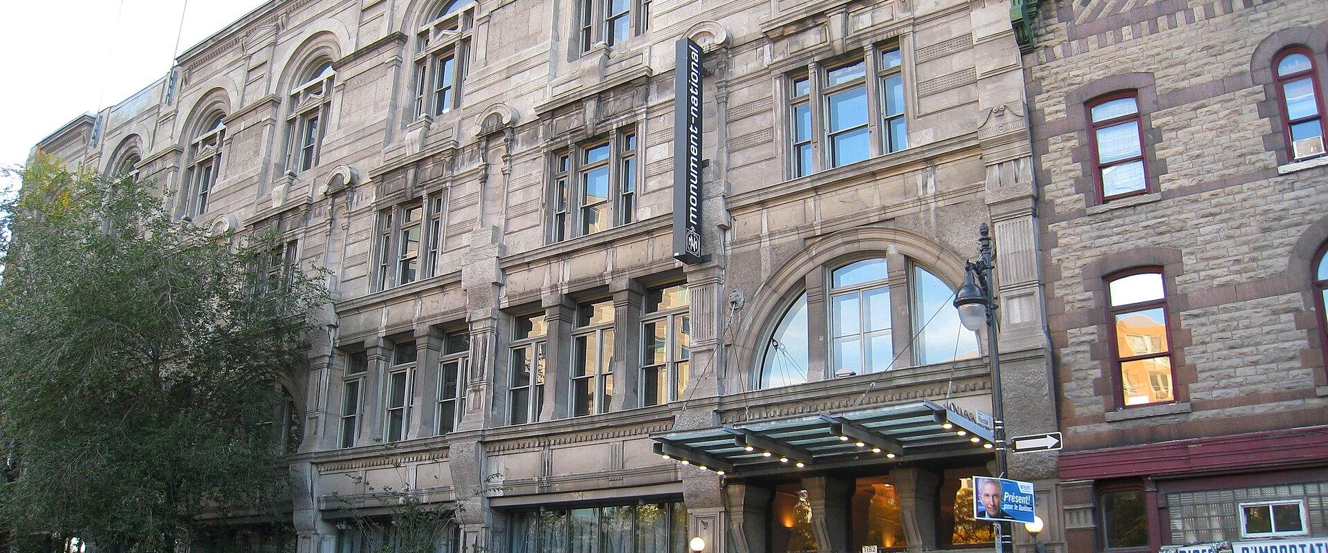A large, ornate stone building on a city street corner featuring a vertical black sign that reads "monument national" and a street-level entrance with a glass canopy.
