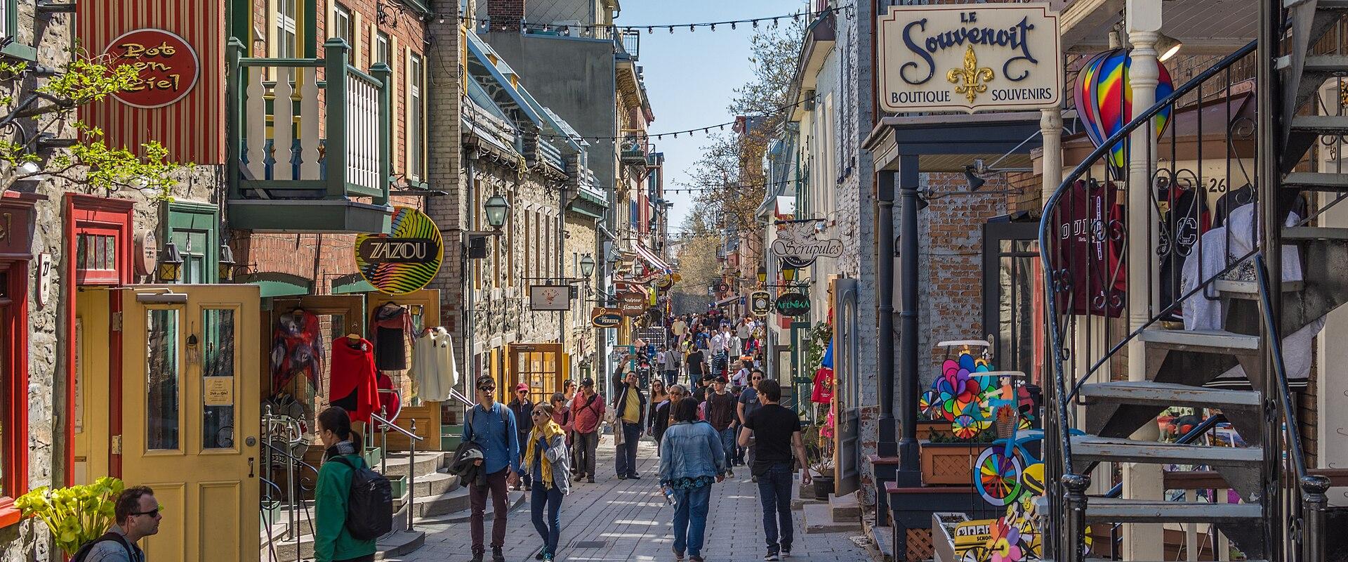A cobblestone pedestrian street lined with historic stone buildings, colorful shopfronts, and hanging signs, including one for "Le Souvenoir" and another for "Bob en Ciel," with pedestrians walking along the path.