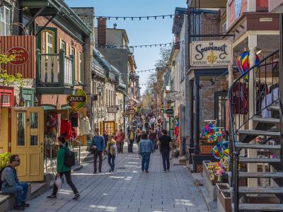 A cobblestone pedestrian street lined with historic stone buildings, colorful shopfronts, and hanging signs, including one for "Le Souvenoir" and another for "Bob en Ciel," with pedestrians walking along the path.