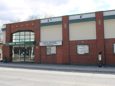 A red brick building with a green awning displaying the text "CINEMA MAGOG" above the entrance, situated on a street corner.