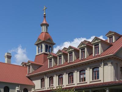 A close-up view of the upper stories and roofline of a large, light-colored institutional building featuring a prominent central cupola topped with a cross, set against a blue sky.