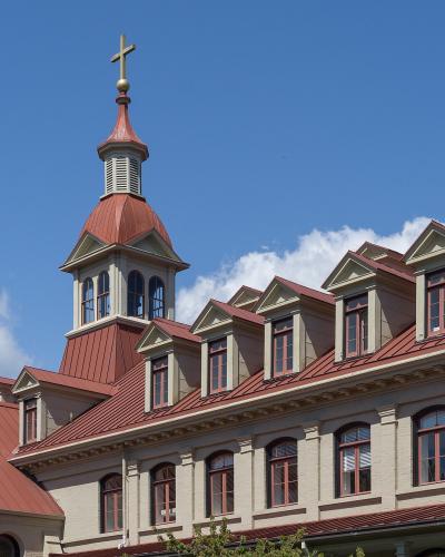 A close-up view of the upper stories and roofline of a large, light-colored institutional building featuring a prominent central cupola topped with a cross, set against a blue sky.